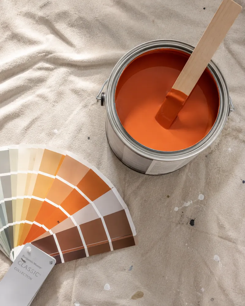Open can of orange paint with a stir stick and a fan deck of color swatches on a drop cloth